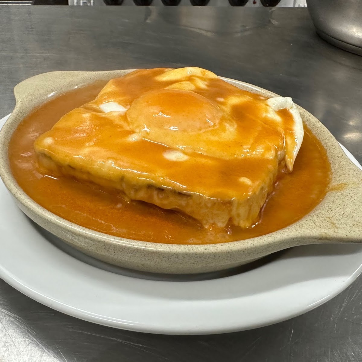 Francesinha em travessa de barro, coberta de queijo derretido e molho alaranjado, com ovo por cima.